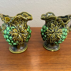 May include: Two ceramic cream pitchers with a grape design. The pitchers are green and brown with a handle and a pedestal base. The grape design is embossed on the front of the pitcher.