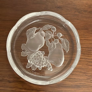 May include: A clear glass coaster featuring an embossed design of pears, grapes, and leaves. The coaster has a slightly scalloped edge.