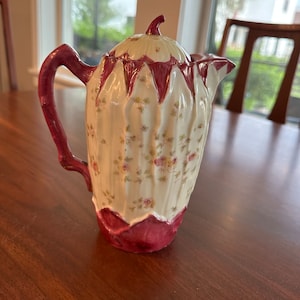 May include: A white ceramic teapot with a pink floral design and a pink lid. The teapot has a handle and a spout.