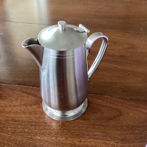 May include: A stainless steel jug with a lid and handle. The jug has a spout for pouring and a rounded base. The handle is curved and attached to the side. The lid has a small knob on top.