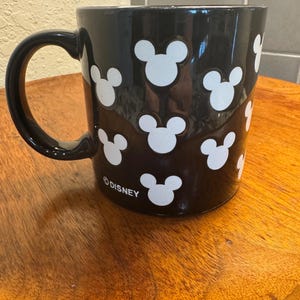 May include: Black ceramic mug with white Mickey Mouse head silhouettes. The mug has a black handle and the word "&copy;DISNEY" printed on the bottom. The mug is sitting on a wooden surface.