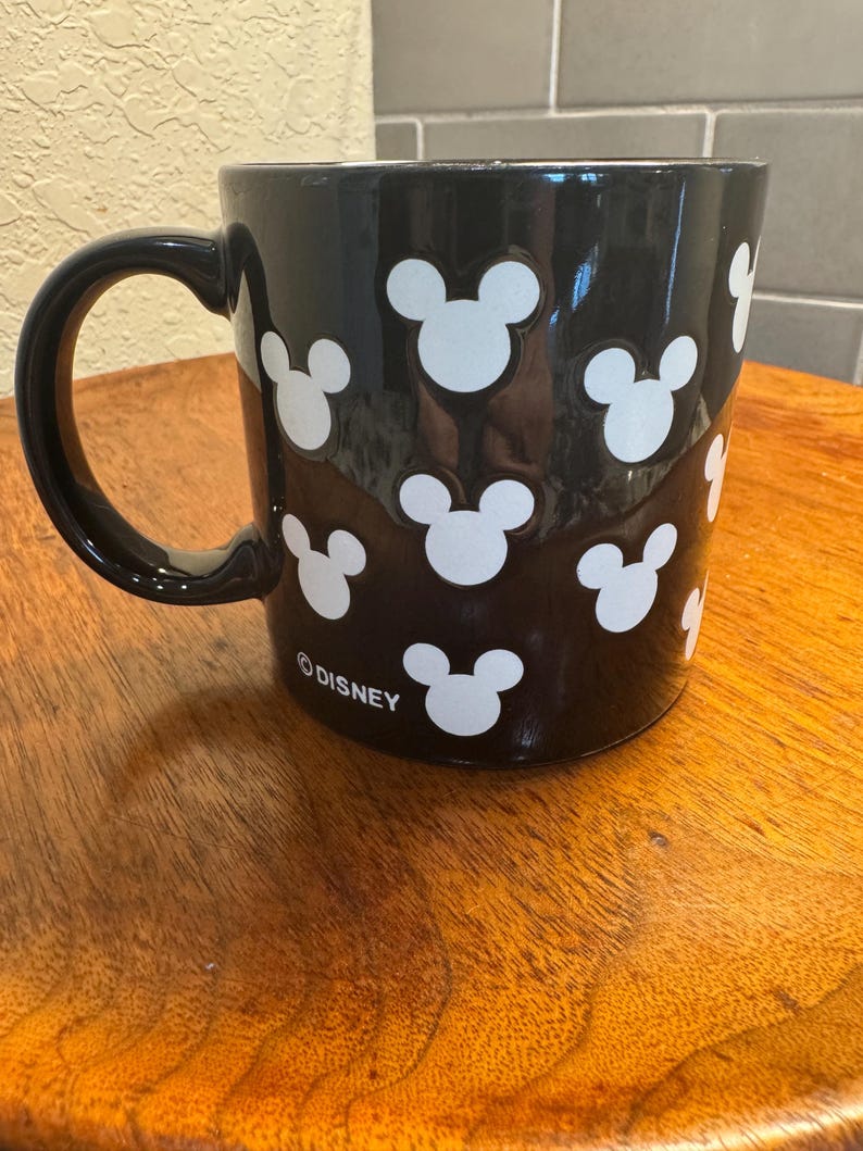 May include: Black ceramic mug with white Mickey Mouse head silhouettes. The mug has a black handle and the word "&copy;DISNEY" printed on the bottom. The mug is sitting on a wooden surface.