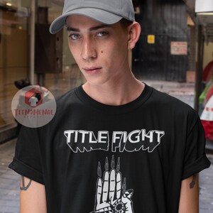 May include: A black t-shirt with a white graphic design of a hand with an eye and the text "TITLE FIGHT". The model is wearing a gray baseball cap.