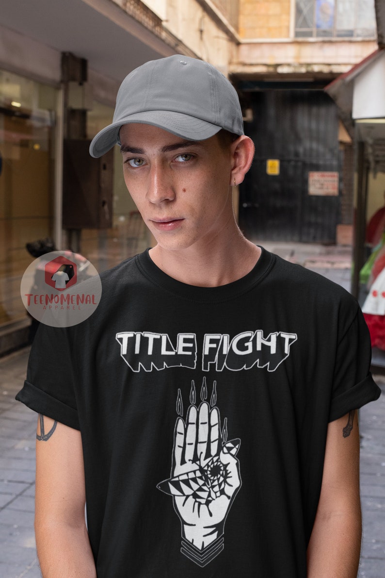 May include: A black t-shirt with a white graphic design of a hand with an eye and the text "TITLE FIGHT". The model is wearing a gray baseball cap.