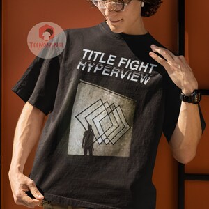 May include: A black t-shirt with the text "TITLE FIGHT HYPERVIEW" printed on the front. The shirt features a graphic of a person standing in front of a series of overlapping squares.