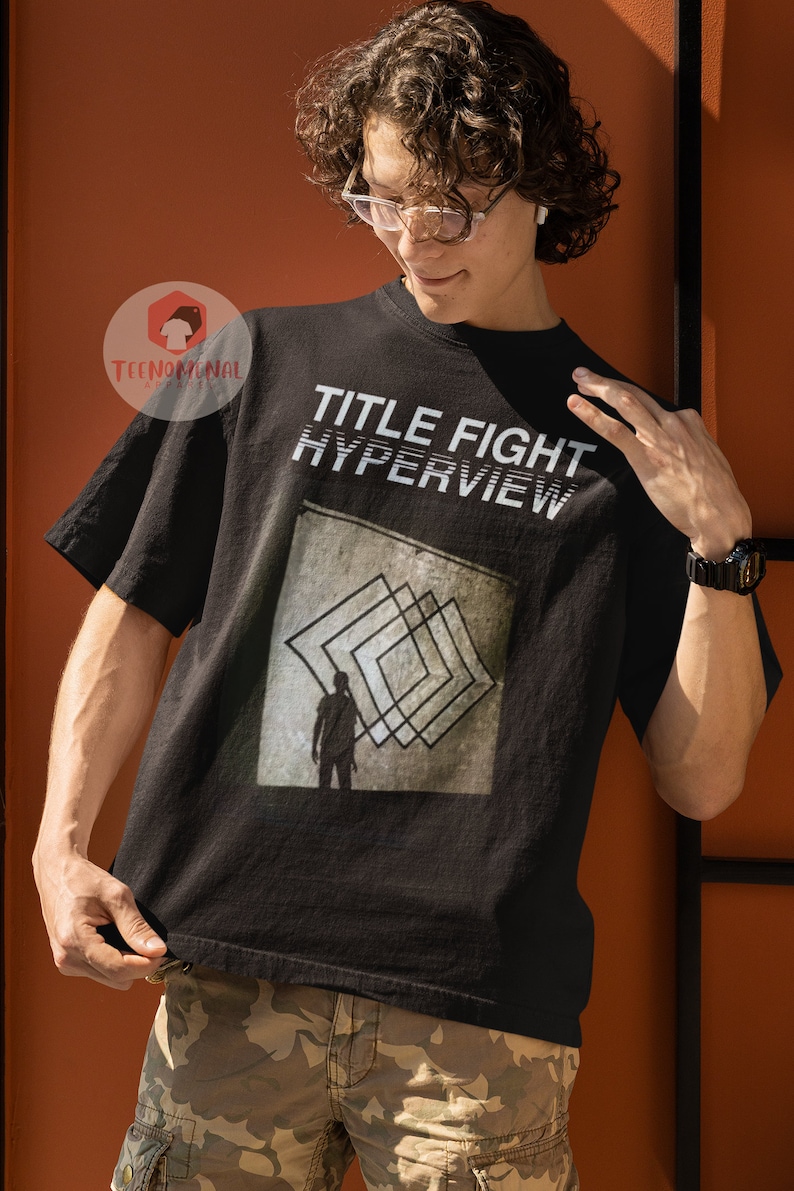 May include: A black t-shirt with the text "TITLE FIGHT HYPERVIEW" printed on the front. The shirt features a graphic of a person standing in front of a series of overlapping squares.