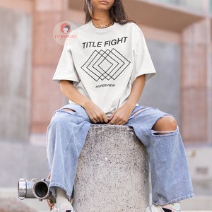 Op de afbeelding: Wit T-shirt met zwarte tekst "TITLE FIGHT" en een geometrisch ontwerp onder de tekst. Het ontwerp bestaat uit overlappende vierkanten en de tekst "HYPERVIEW" staat onder het ontwerp.