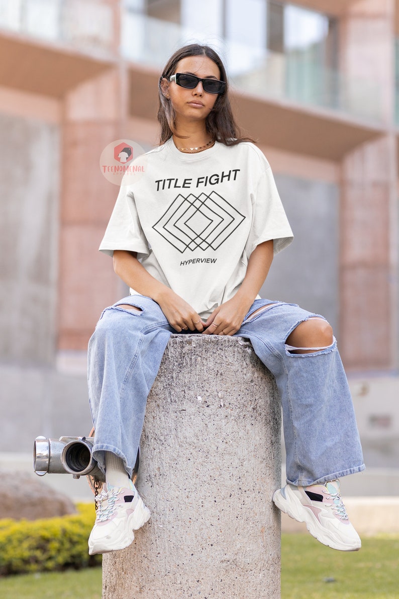 Op de afbeelding: Wit T-shirt met zwarte tekst "TITLE FIGHT" en een geometrisch ontwerp onder de tekst. Het ontwerp bestaat uit overlappende vierkanten en de tekst "HYPERVIEW" staat onder het ontwerp.