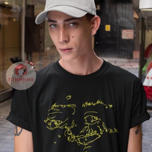 Puede incluir: Una camiseta negra con un dibujo de l&iacute;nea amarilla de una cara y el texto "Mac DeMarco" en amarillo.