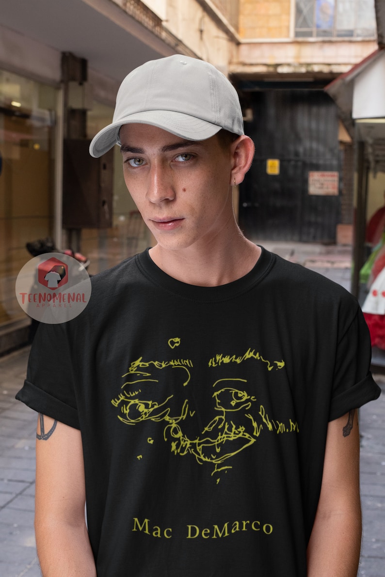 Puede incluir: Una camiseta negra con un dibujo de l&iacute;nea amarilla de una cara y el texto "Mac DeMarco" en amarillo.