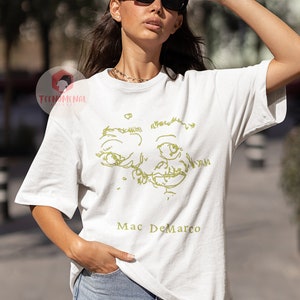 Puede incluir: Camiseta blanca con un dibujo de l&iacute;nea verde de una cara y el texto "Mac DeMarco" impreso en la parte delantera.