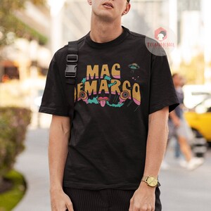 May include: Black t-shirt with the text "MAC DEMARCO" in a colourful, retro font, and a UFO graphic. The shirt is worn by a person wearing sunglasses and a black cap. A gold watch is on the person's wrist.