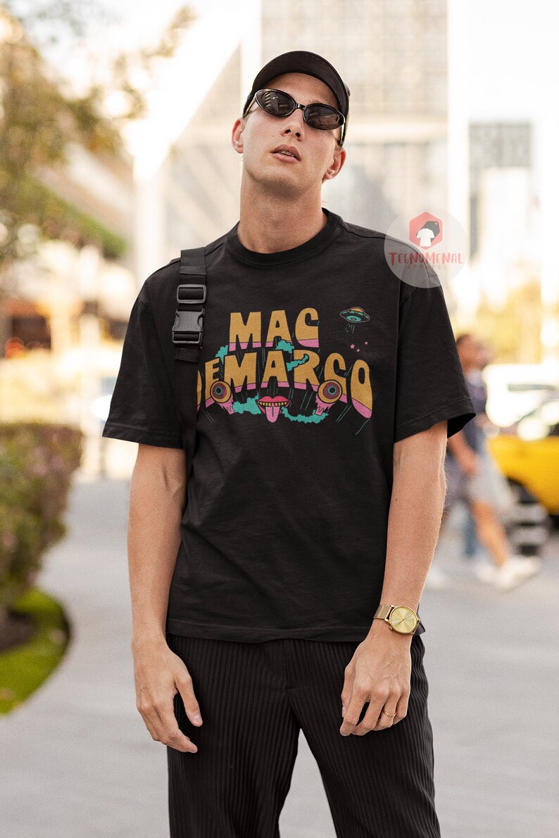 May include: Black t-shirt with the text "MAC DEMARCO" in a colourful, retro font, and a UFO graphic. The shirt is worn by a person wearing sunglasses and a black cap. A gold watch is on the person's wrist.