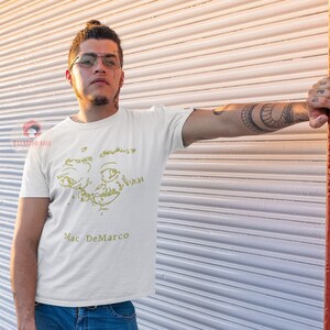 Puede incluir: Una camiseta blanca con un dibujo de l&iacute;nea amarilla de una cara y el texto "Mac DeMarco" impreso en la parte delantera.