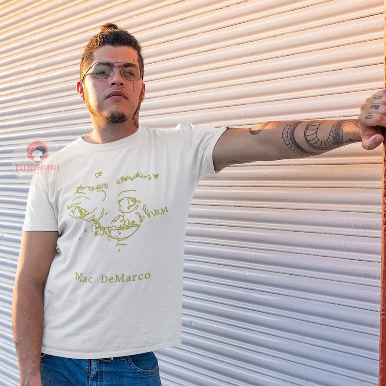 Puede incluir: Una camiseta blanca con un dibujo de l&iacute;nea amarilla de una cara y el texto "Mac DeMarco" impreso en la parte delantera.