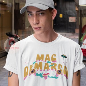 May include: White t-shirt with the text "MAC DEMARCO" in gold, pink, and teal. The design includes cartoon eyes, a mouth, and a flying saucer. The person is wearing a grey baseball cap.