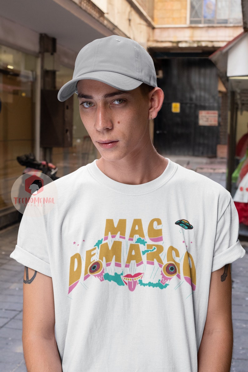 May include: White t-shirt with the text "MAC DEMARCO" in gold, pink, and teal. The design includes cartoon eyes, a mouth, and a flying saucer. The person is wearing a grey baseball cap.