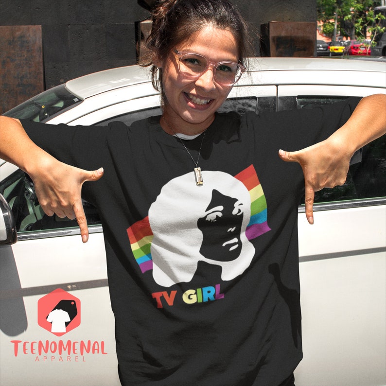 May include: A black t-shirt featuring a white silhouette of a woman's face with a rainbow stripe behind it. The text "TV GIRL" is printed below the image.