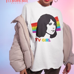 May include: White t-shirt featuring a black and white illustration of a woman with a rainbow behind her and the text "TV GIRL" underneath.