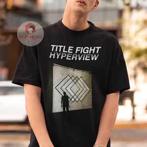 May include: Black t-shirt with white text that reads "TITLE FIGHT HYPERVIEW". The shirt features a graphic of a person standing in front of a wall with a geometric pattern.