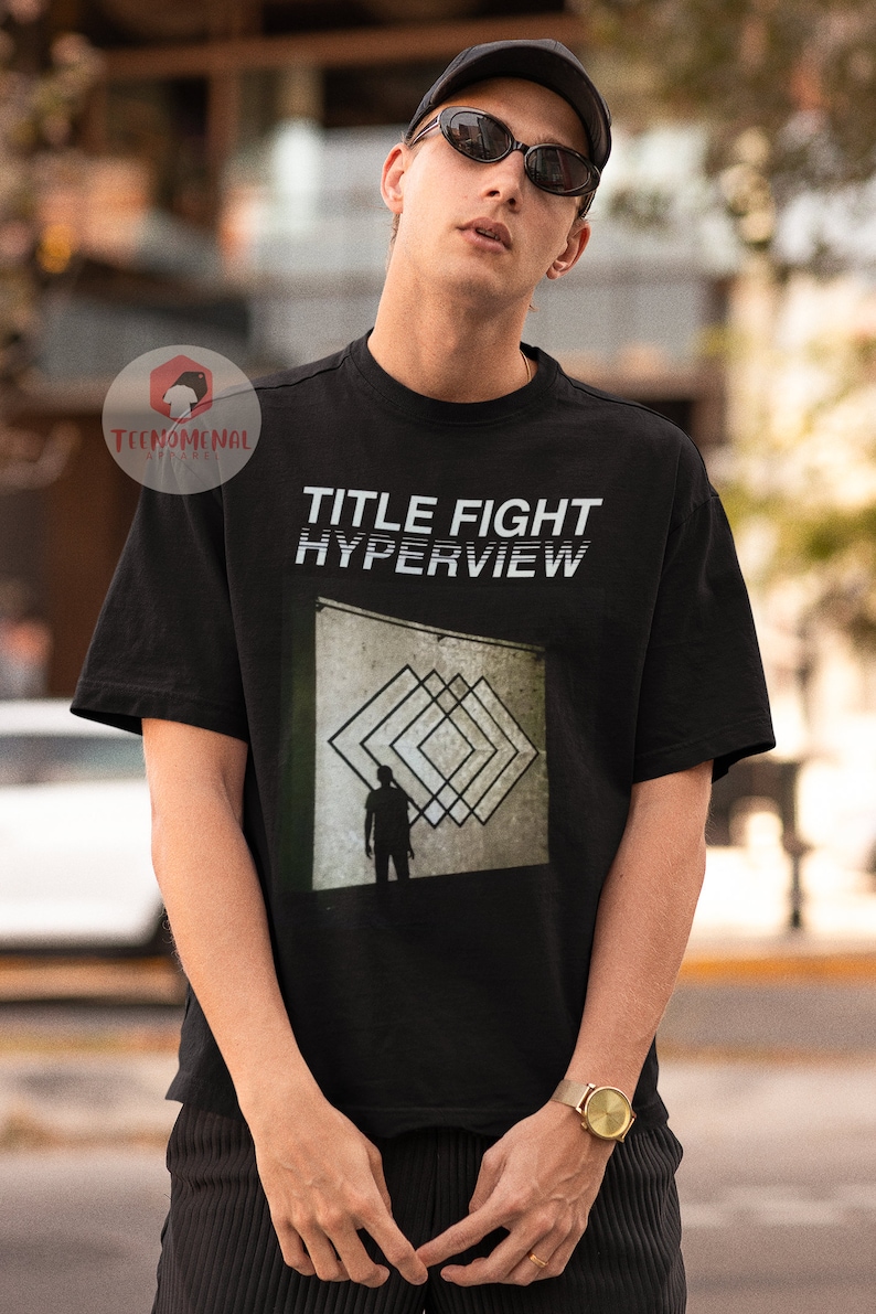 May include: Black t-shirt with white text that reads "TITLE FIGHT HYPERVIEW". The shirt features a graphic of a person standing in front of a wall with a geometric pattern.