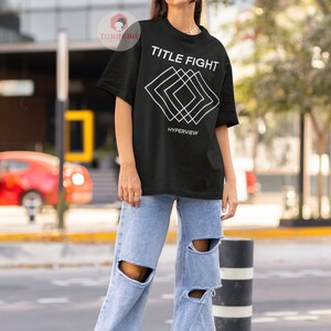 Op de afbeelding: Zwart T-shirt met een wit grafisch ontwerp met de tekst "TITLE FIGHT" en "HYPERVIEW". De grafiek is een reeks overlappende witte vierkanten.