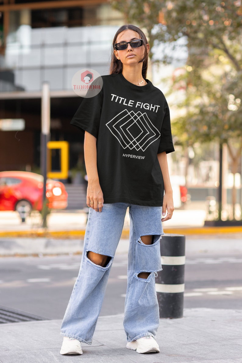 Op de afbeelding: Zwart T-shirt met een wit grafisch ontwerp met de tekst "TITLE FIGHT" en "HYPERVIEW". De grafiek is een reeks overlappende witte vierkanten.