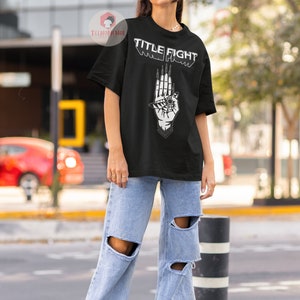 May include: A black t-shirt with a white graphic of a hand with an eye and the text "TITLE FIGHT" printed on the front.