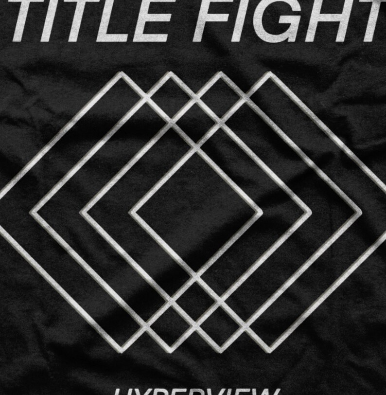 Op de afbeelding: Zwart T-shirt met een wit grafisch ontwerp van overlappende vierkanten en de tekst "TITLE FIGHT" en "HYPERVIEW".