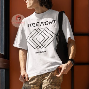 Op de afbeelding: Een wit T-shirt met zwarte tekst die "TITLE FIGHT" zegt en een geometrisch ontwerp eronder. De tekst "HYPERVIEW" staat onder het ontwerp.