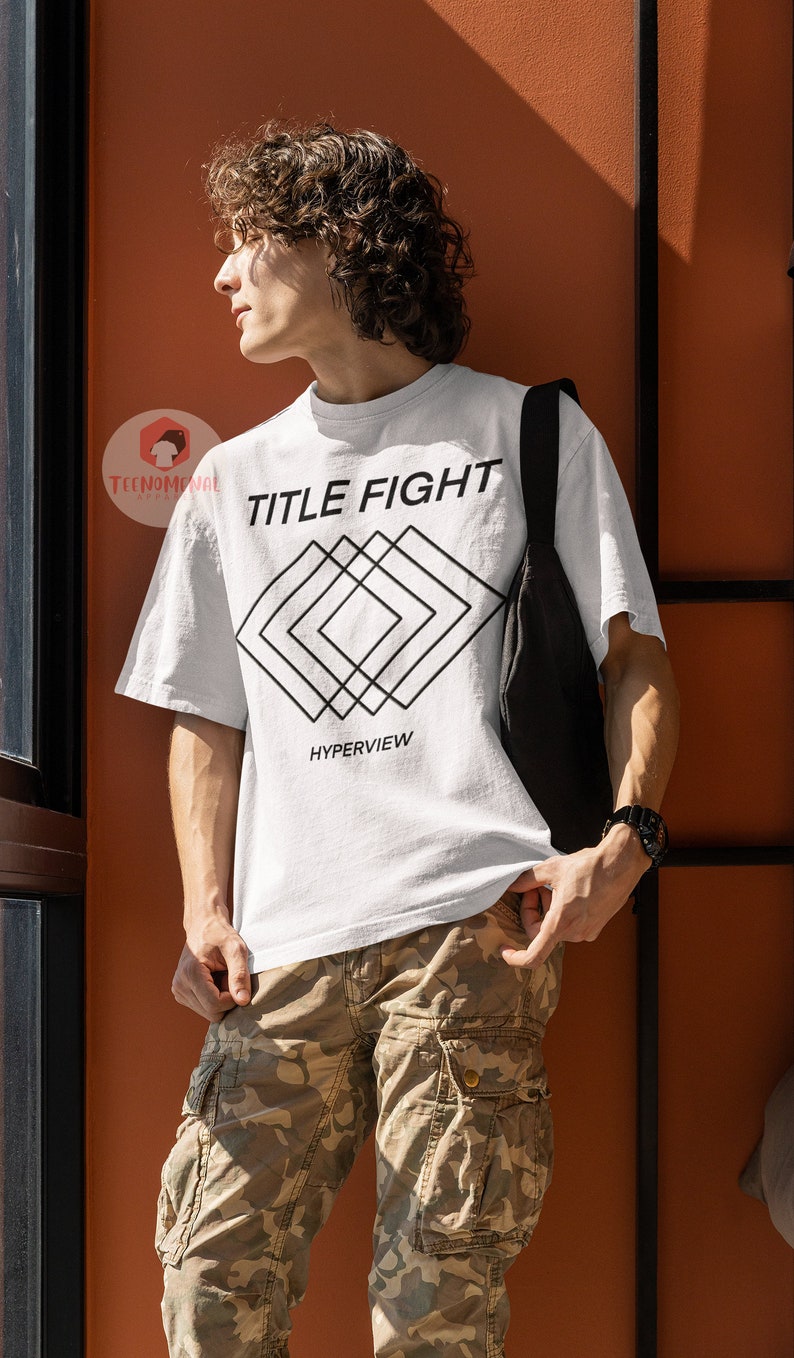 Op de afbeelding: Een wit T-shirt met zwarte tekst die "TITLE FIGHT" zegt en een geometrisch ontwerp eronder. De tekst "HYPERVIEW" staat onder het ontwerp.