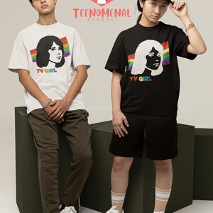 May include: Two people wearing white and black t-shirts with a graphic of a woman's face and the text "TV GIRL". The t-shirts have a rainbow stripe design. The people are standing on a green platform. The logo "TEENOMENAL APPAREL" is visible in the top centre of the image.