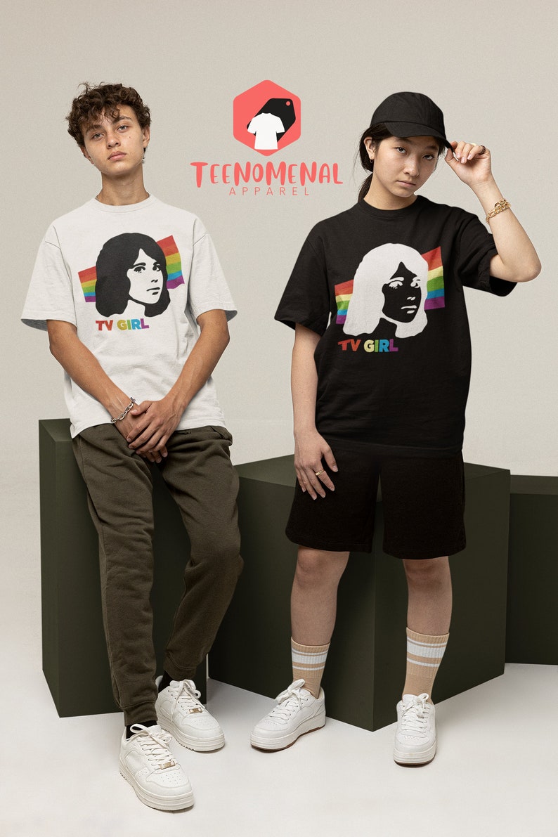 May include: Two people wearing white and black t-shirts with a graphic of a woman's face and the text "TV GIRL". The t-shirts have a rainbow stripe design. The people are standing on a green platform. The logo "TEENOMENAL APPAREL" is visible in the top centre of the image.