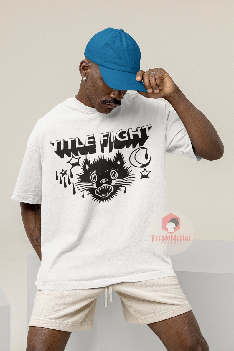 Może przedstawiać: Biały t-shirt z czarnym nadrukiem kota z tekstem "TITLE FIGHT" nad nim. Kot ma groźny wyraz twarzy i jest otoczony gwiazdami, rogalem księżyca i kapiącą krwią.