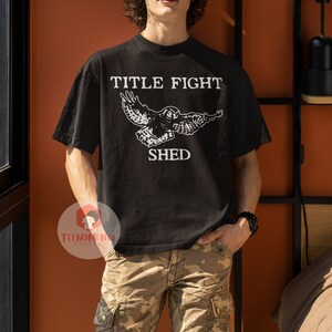 Puede incluir: Una camiseta negra con un gr&aacute;fico blanco de un b&uacute;ho volando con el texto "TITLE FIGHT SHED" impreso en la parte delantera.
