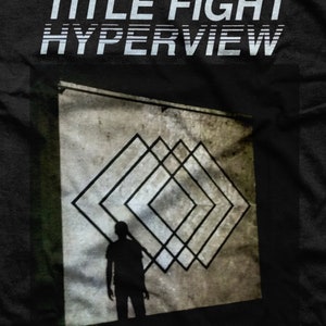 May include: A black and white image of a person standing in front of a wall with a geometric pattern of overlapping squares. The text "TITLE FIGHT HYPERVIEW" is at the top of the image.