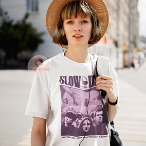 May include: A white t-shirt featuring a purple graphic print of a band called "Slowdive". The graphic depicts a band member playing a guitar and the band's name in large letters. The text "Just for a Day - Souvlaki - Pygmalion - Slowdive" is printed beneath the graphic.