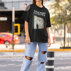 May include: A black t-shirt with the text "TITLE FIGHT HYPERVIEW" and a graphic of a person standing in front of a geometric design. The person is wearing a black shirt and black pants. The graphic is in shades of gray and white.