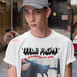 Pu&ograve; includere: Una maglietta bianca con una stampa grafica nera e rossa di un gatto e il testo "TITLE FIGHT KINGSTON PA USA". La persona che indossa la maglietta indossa anche un berretto da baseball grigio.