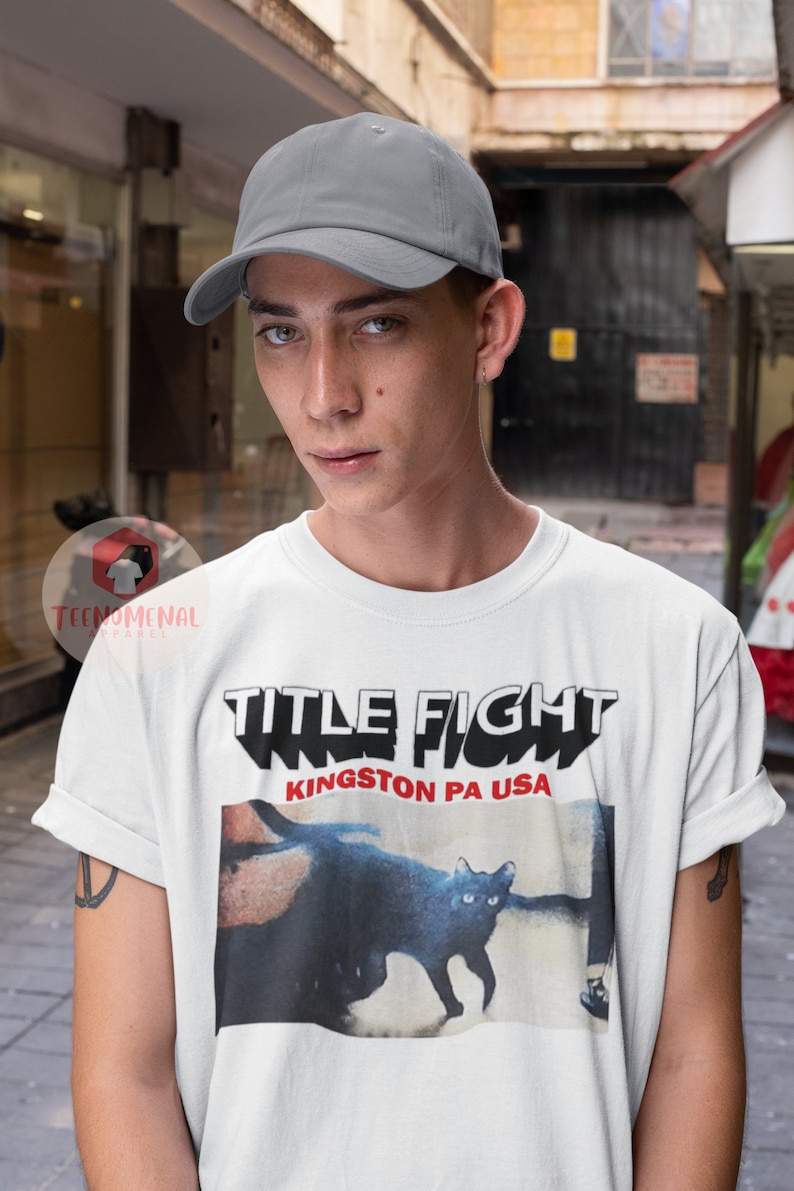 Pu&ograve; includere: Una maglietta bianca con una stampa grafica nera e rossa di un gatto e il testo "TITLE FIGHT KINGSTON PA USA". La persona che indossa la maglietta indossa anche un berretto da baseball grigio.