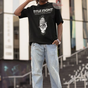 May include: A black t-shirt with a white graphic of a hand with an eye and the text "TITLE FIGHT" printed on the front.