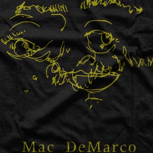 Puede incluir: Una camiseta negra con un dibujo de l&iacute;nea amarilla de una cara y el texto "Mac DeMarco" en amarillo.