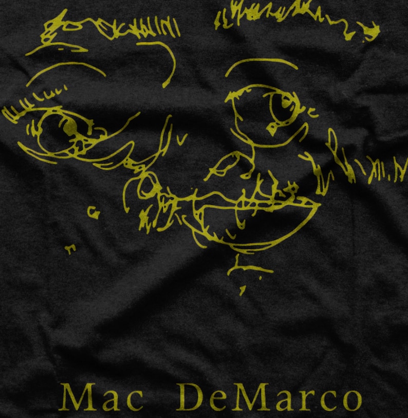 Puede incluir: Una camiseta negra con un dibujo de l&iacute;nea amarilla de una cara y el texto "Mac DeMarco" en amarillo.
