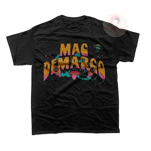 May include: Black t-shirt with the text "MAC DEMARCO" in gold and pink, featuring a graphic of a mouth, eyes, and a UFO. The shirt is a crew neck and has short sleeves.