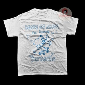 May include: White t-shirt with a blue graphic of Snoopy wearing headphones and riding a skateboard. The shirt has the text "LEAVE ME ALONE, I'm listening to Turnstile" in blue. A fun, casual tee.