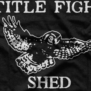 Puede incluir: Camiseta negra con un gr&aacute;fico blanco de un p&aacute;jaro en vuelo y el texto "TITLE FIGHT SHED" debajo.