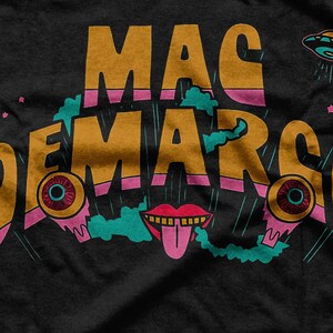 May include: Black t-shirt featuring the text "MAC DEMARCO" in gold lettering with pink and teal details. The design incorporates cartoon eyes, a tongue, and a UFO.
