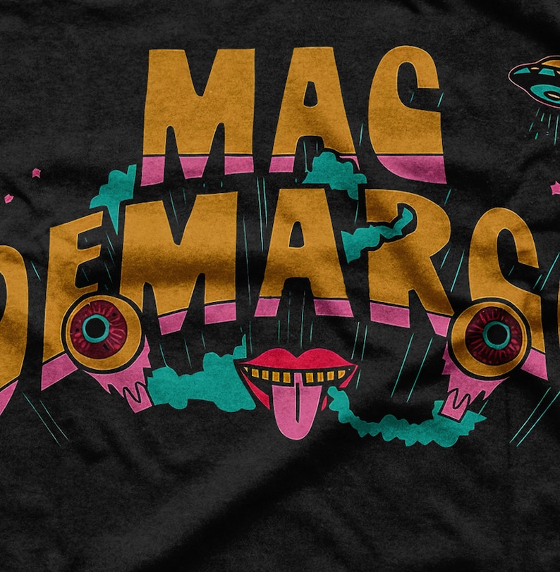 May include: Black t-shirt featuring the text "MAC DEMARCO" in gold lettering with pink and teal details. The design incorporates cartoon eyes, a tongue, and a UFO.