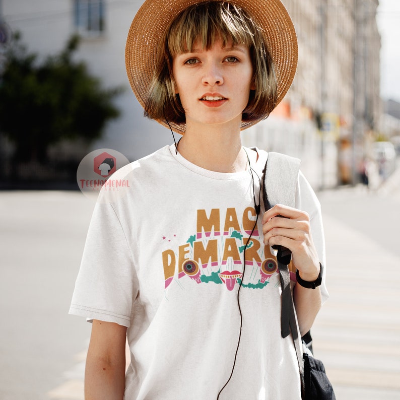 May include: A person wearing a white t-shirt with the text "MAC DEMARCO" in brown and pink lettering. The shirt features a colourful graphic design with a tongue, eyes, and musical elements. The person is wearing a straw hat and carrying a black bag.