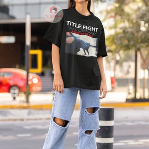 Pu&ograve; includere: Maglietta nera con una stampa grafica di un gatto nero e il testo "TITLE FIGHT" e "KINGSTON PA USA".